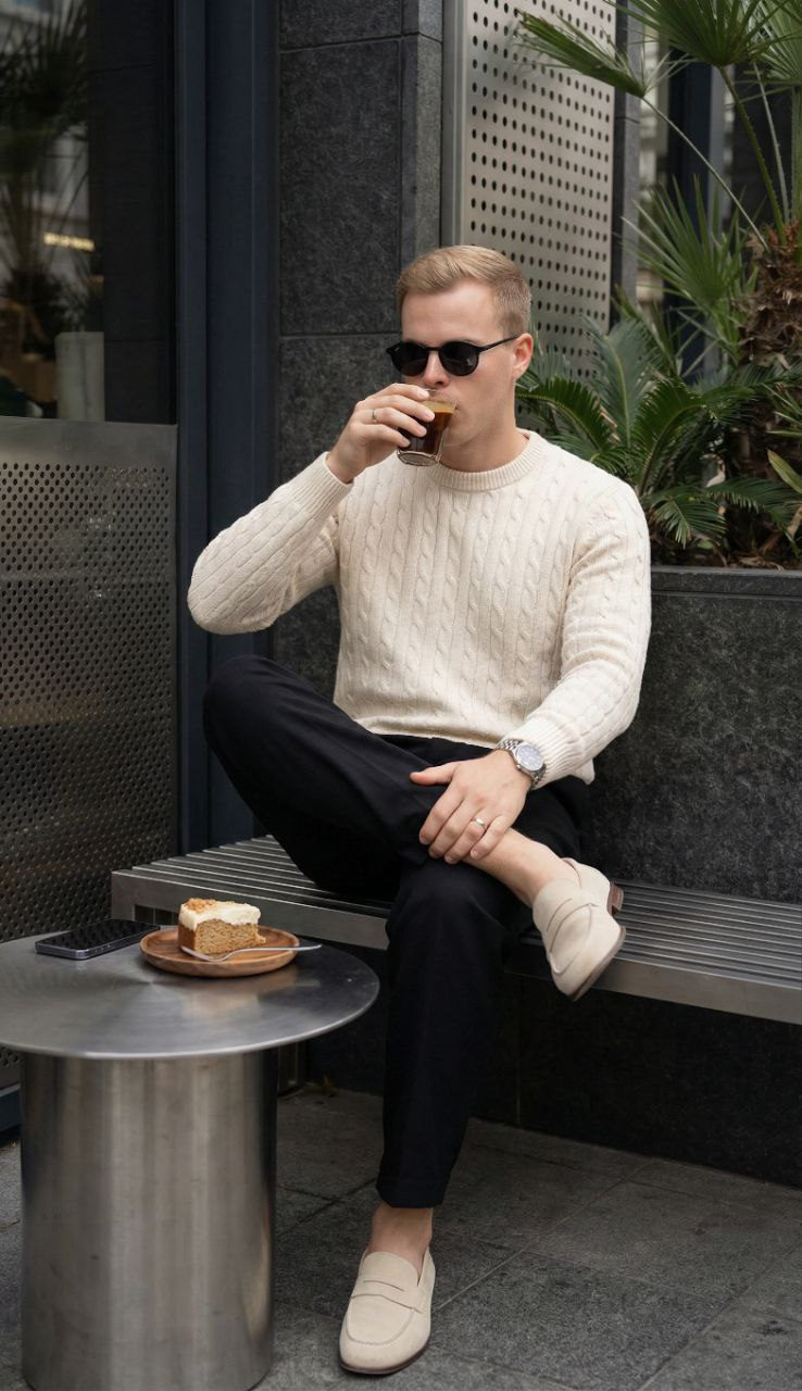 A man in a cream sweater sits on a metal bench, sipping coffee at a stylish urban café.