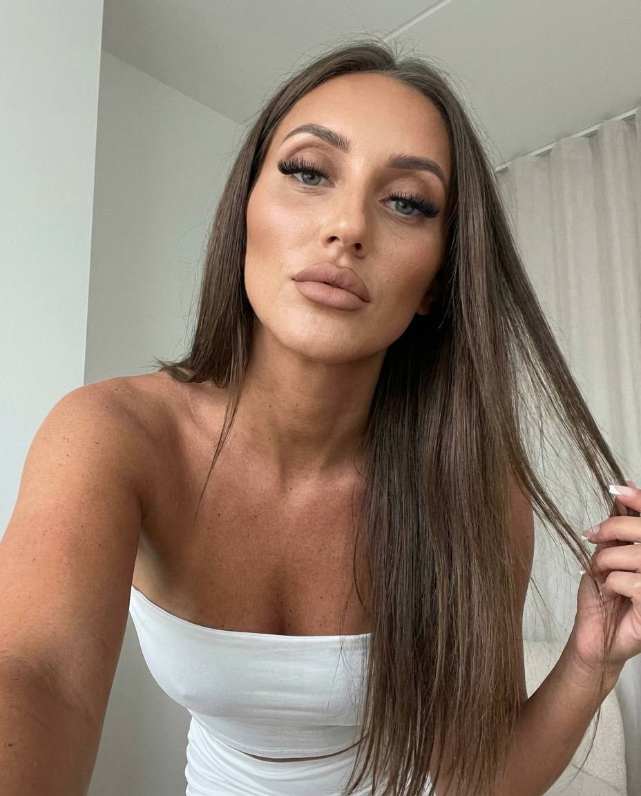 A girl in a white outfit takes a selfie indoors with soft diffused light, leaning forward with a confident expression.