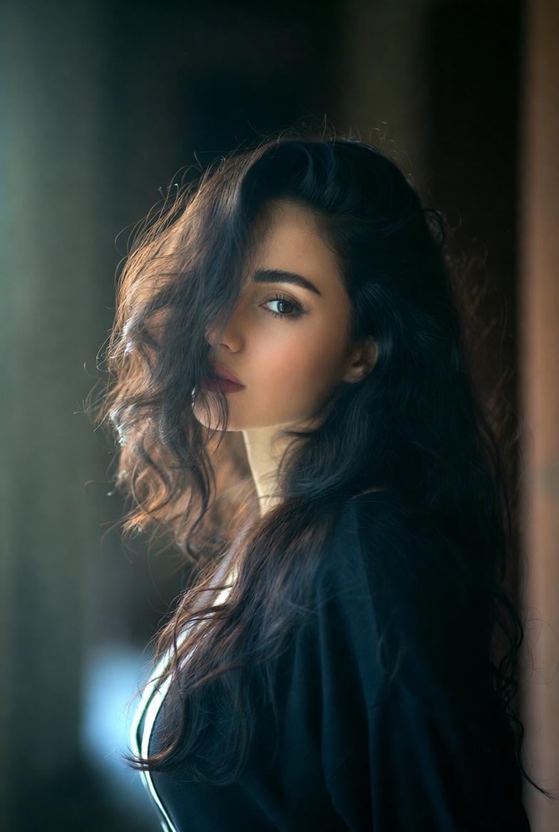 A medium close-up shot of a person with dark, wavy hair over their shoulder, softly illuminated by diffused natural light.