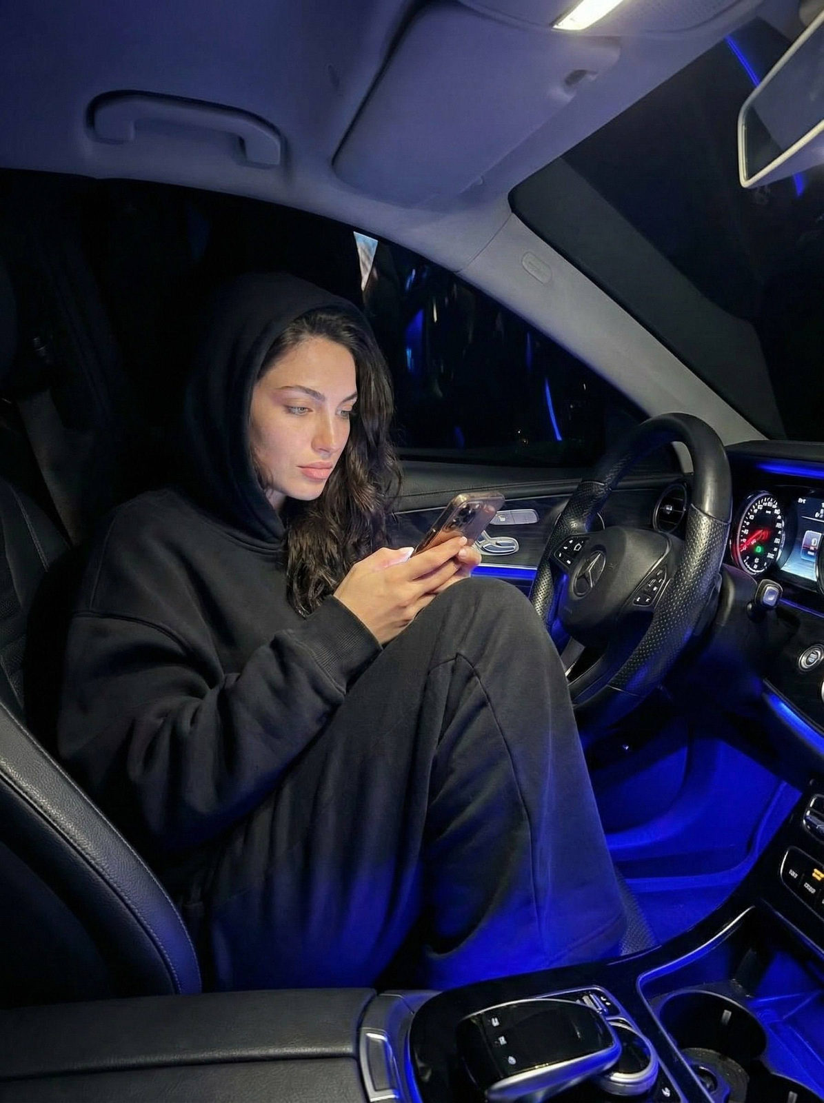 Photorealistic image of a girl in a luxury car at night, holding a smartphone, illuminated by neon blue light.