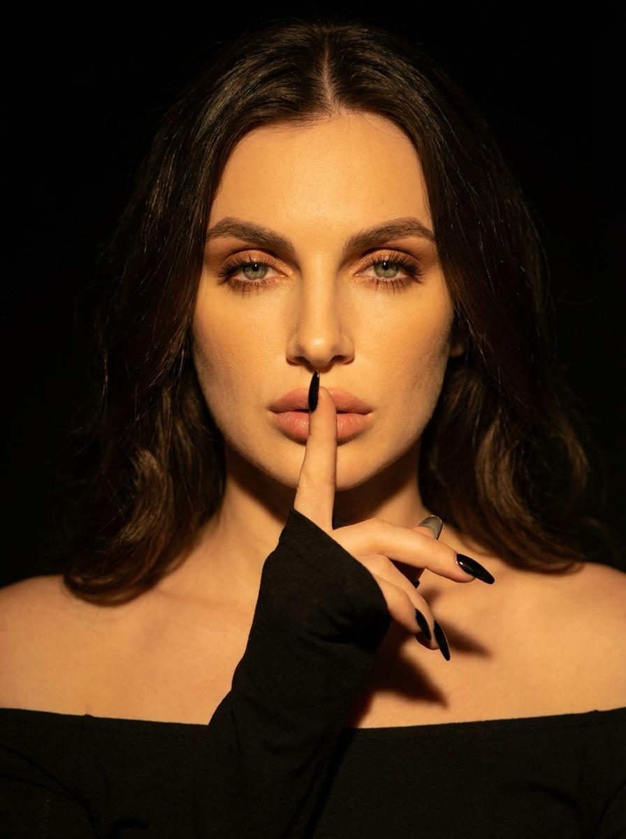 A woman in a dark room, her finger to her lips in silence, wearing a black dress, with soft waves framing her face and a warm golden glow.