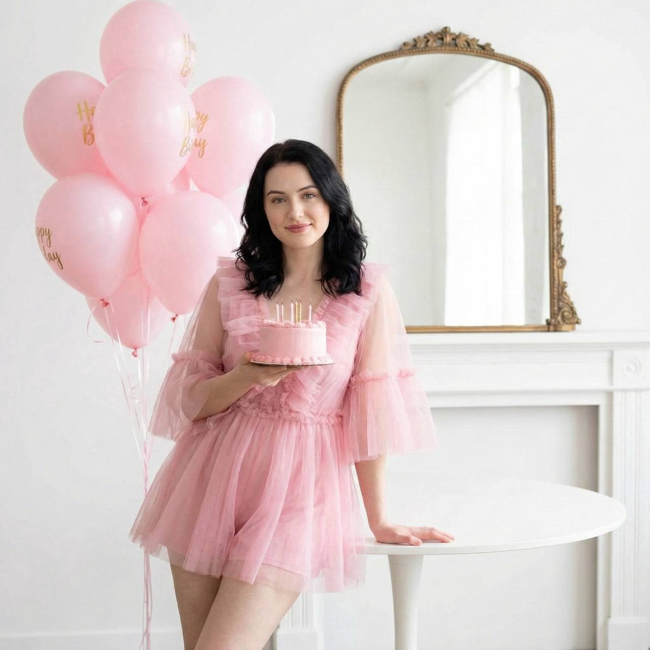 A woman in a pink tulle jumpsuit holding a pink birthday cake with lit candles in a bright room decorated with light pink balloons.
