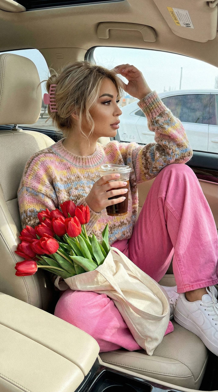 Close-up of a girl in a light-colored car interior on a sunny day, wearing bright pink jeans and a patterned sweater while holding iced coffee.
