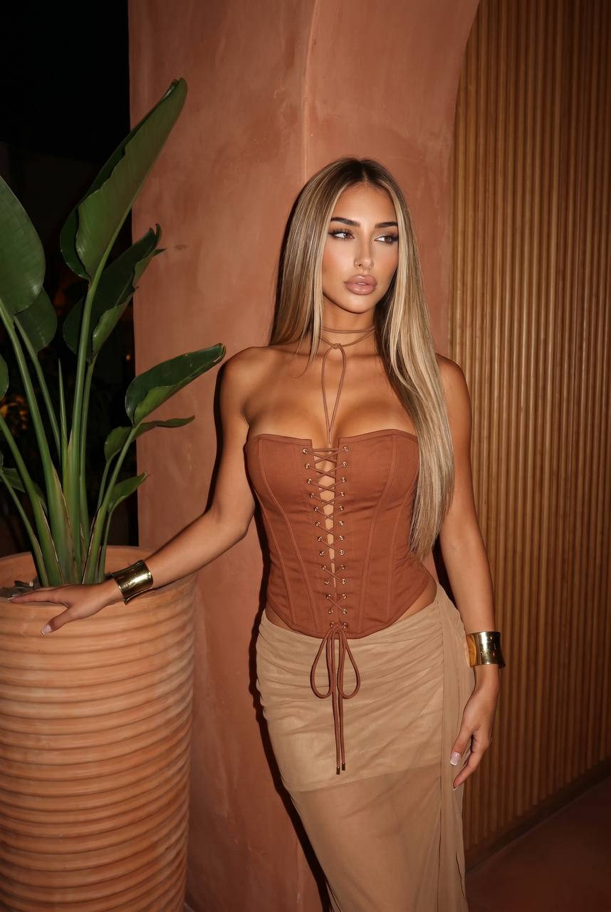 Woman wearing a rust corset and tan skirt in a warm-toned interior beside a tall green plant, calm expression and sleek hair.