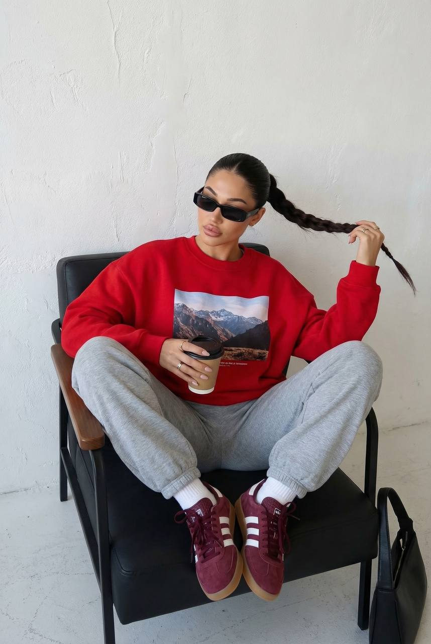 Portrait of a confident woman in a red sweatshirt sitting in a dark armchair, holding a coffee cup, with a relaxed pose.