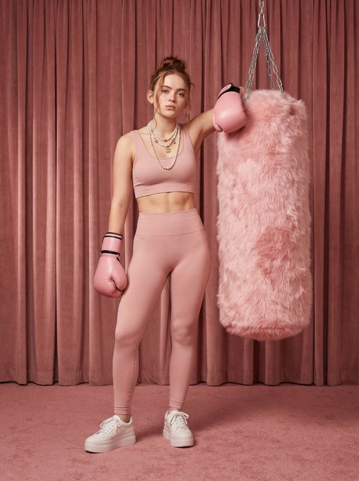 A full-body shot of a model in a dusty rose athletic set, wearing pale pink boxing gloves and layered pearl necklaces, set against a pink velvet backdrop.