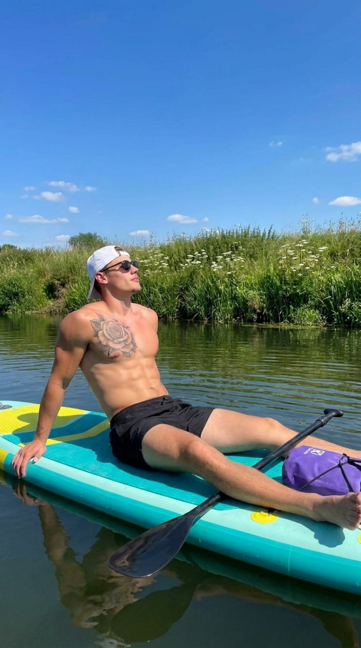 A man with short brown hair and sunglasses relaxing on a turquoise inflatable surfboard on calm green river water.
