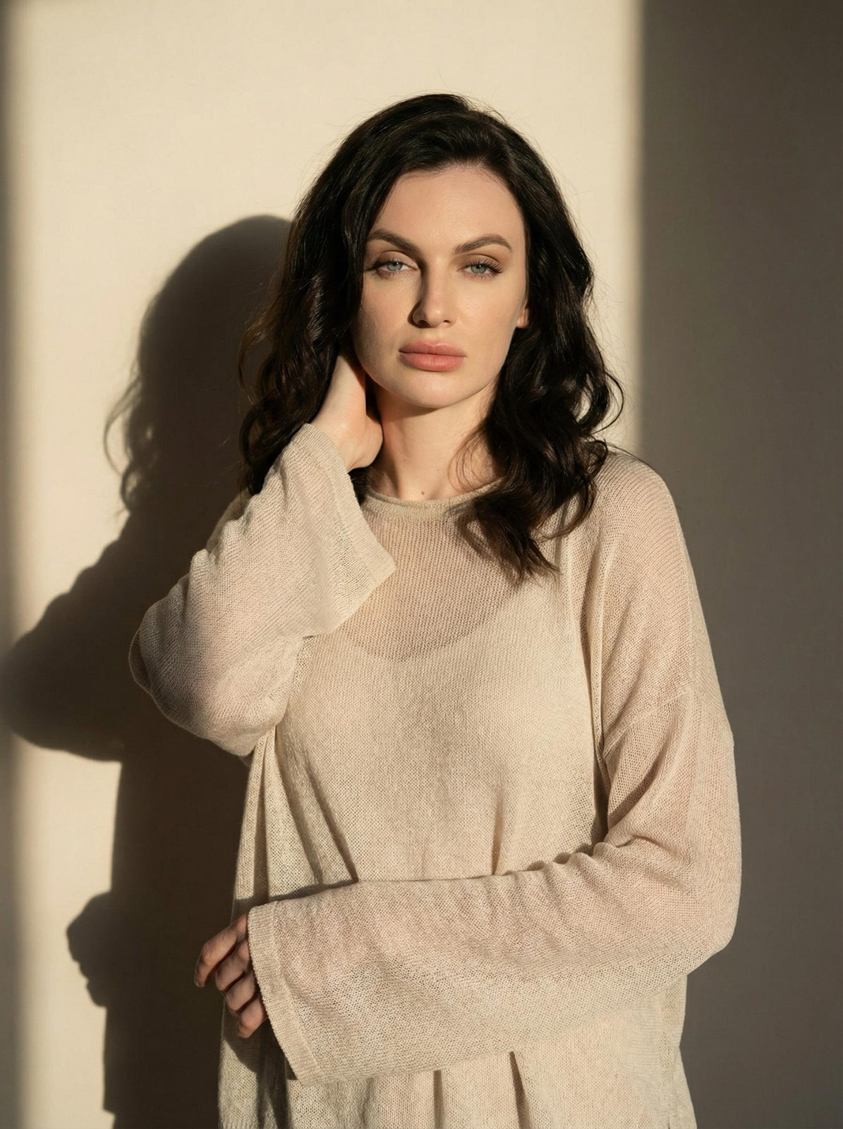 A woman in a semi-sheer knit sweater posed against a light wall, showcasing contrast between light and shadow.