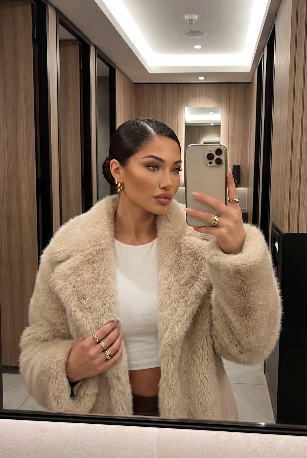 Ultra-realistic fashion mirror selfie highlighting a woman with a sleek low bun, wearing a light beige faux fur coat in a high-end restroom.