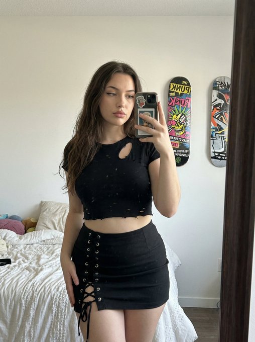A woman in a black, distressed crop top and mini skirt taking a mirror selfie in a softly lit bedroom.