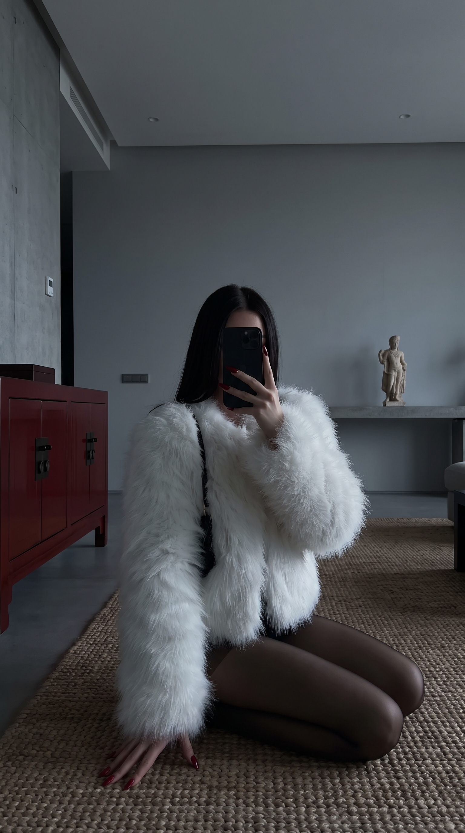 A young woman kneeling on a textured mat with red nails, holding a black iPhone while wearing a fluffy white jacket.