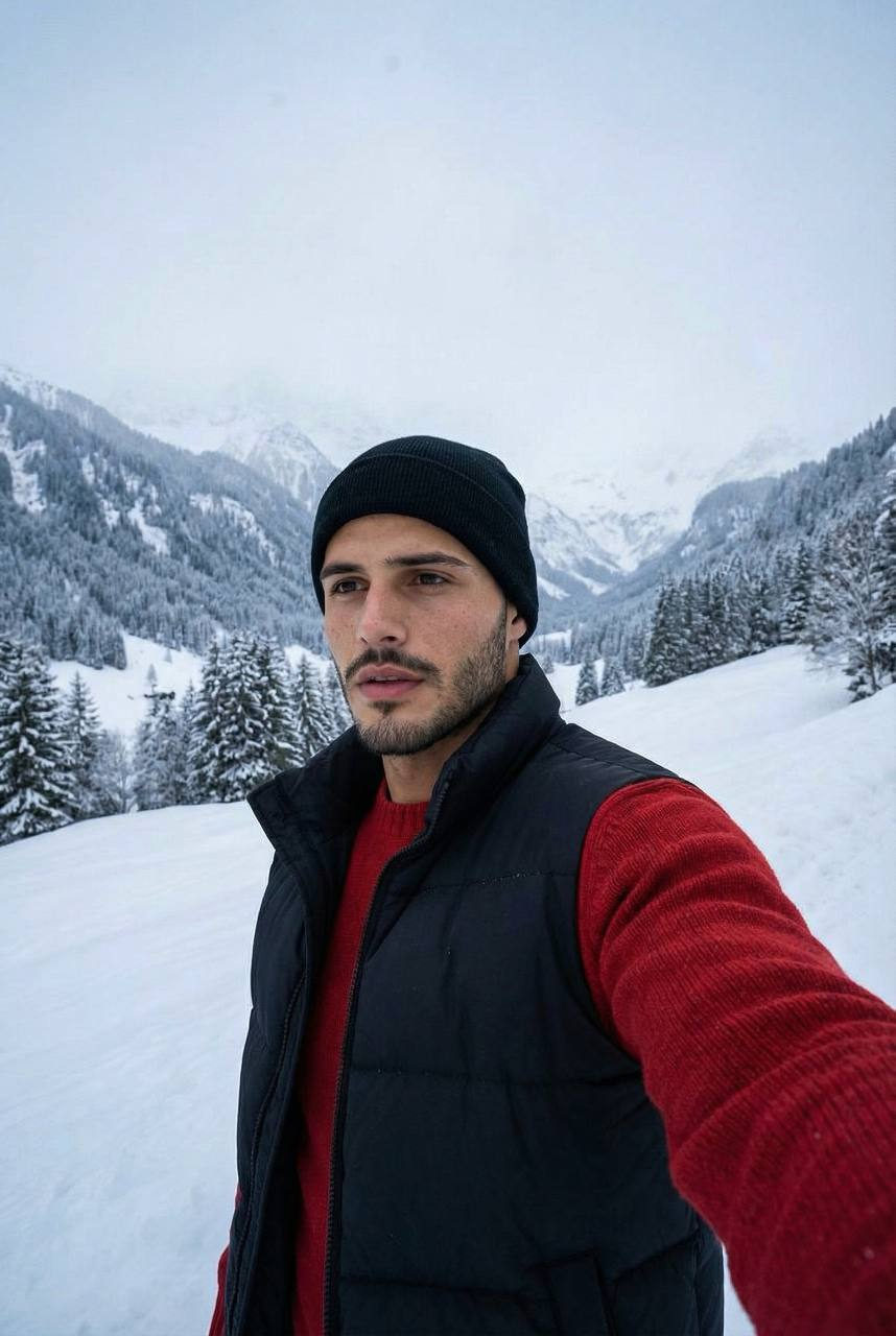 Handheld selfie of a man in a winter landscape wearing a red sweater and black vest, snow-covered mountains in the background.
