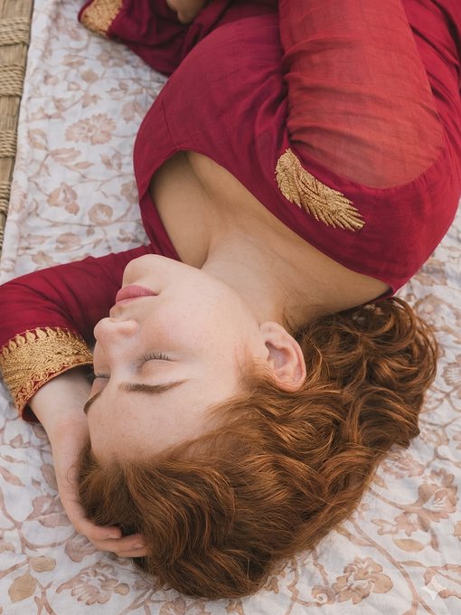 Close-up of Sadie Sink resting on a woven bed with curly hair and a maroon tunic, serene expression in natural light.