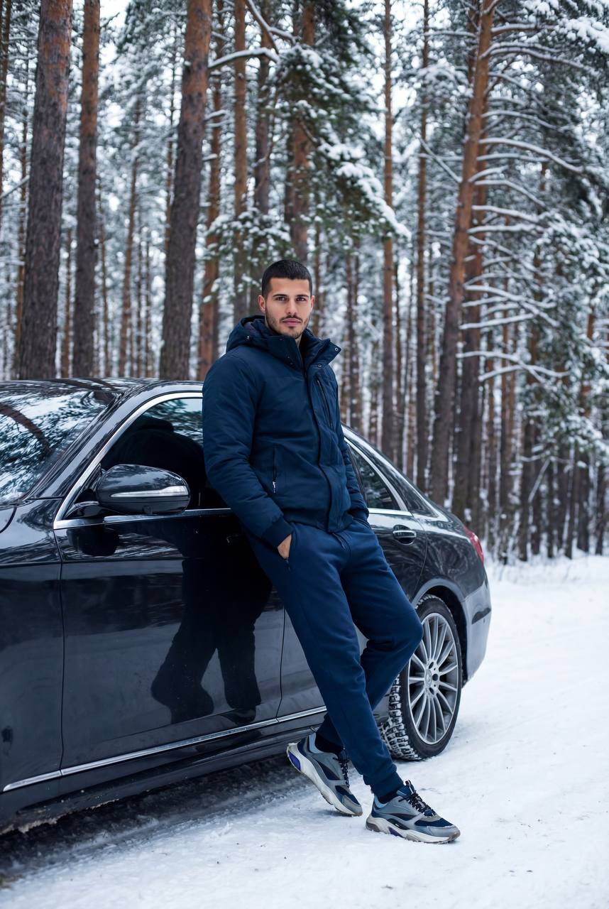 A person in a dark blue winter tracksuit leans against a luxury sedan in a snowy forest, showcasing a calm and confident demeanor.