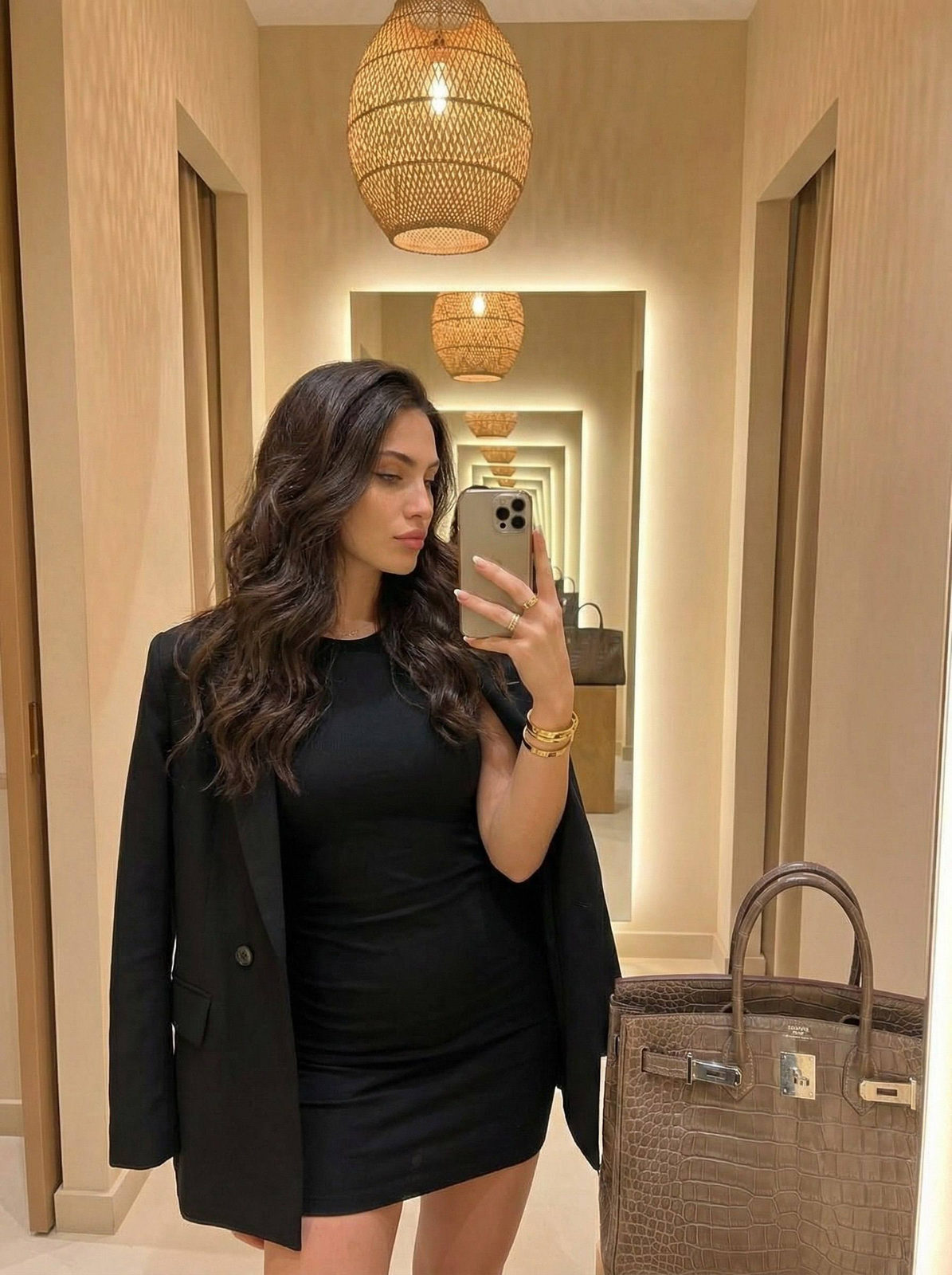 A luxurious cozy mirror selfie featuring a woman in a tight black mini dress and elegant blazer, posed with a phone.