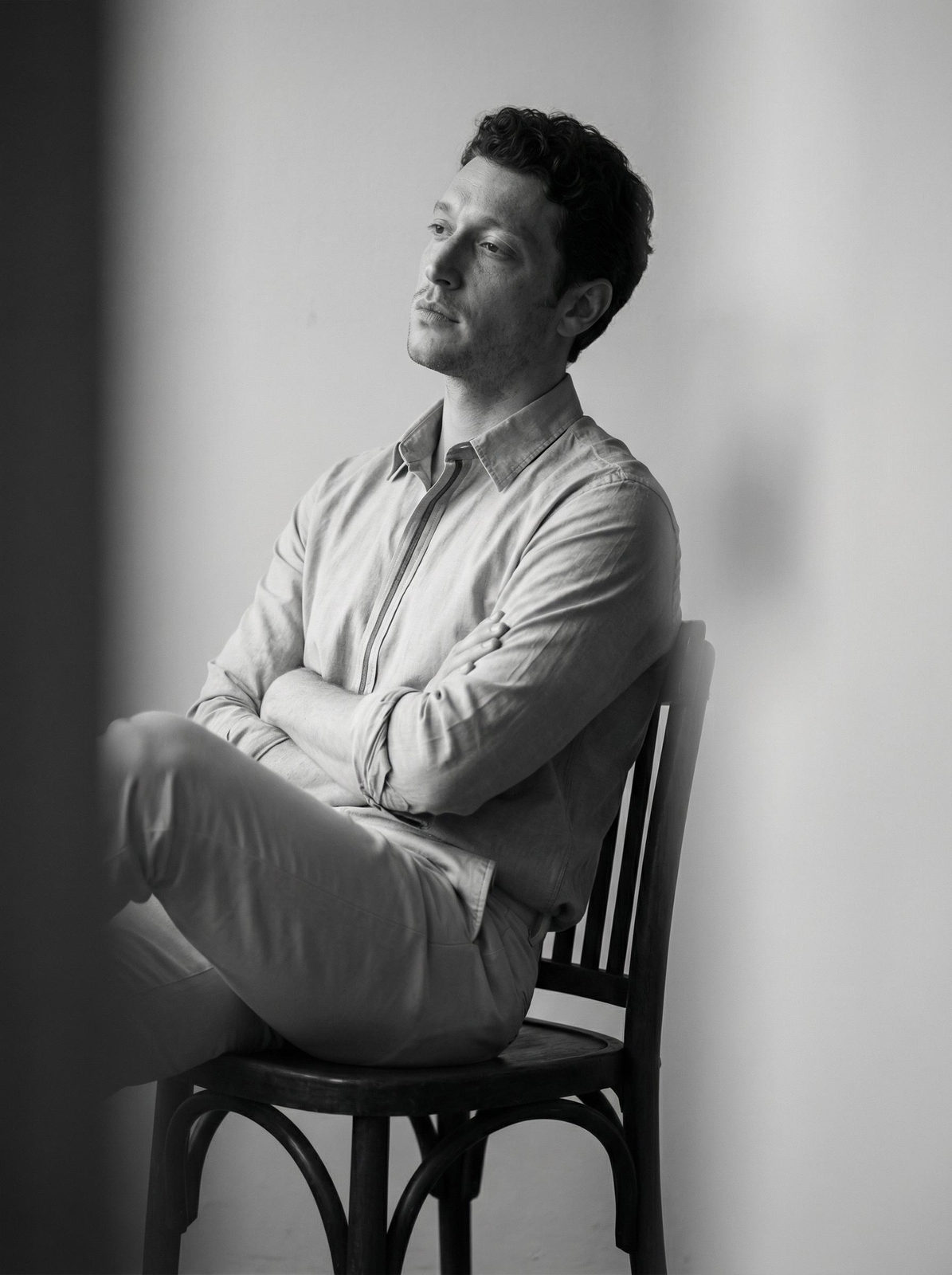 Moody monochrome portrait of a man seated on a dark wooden chair with blurred gray foreground.