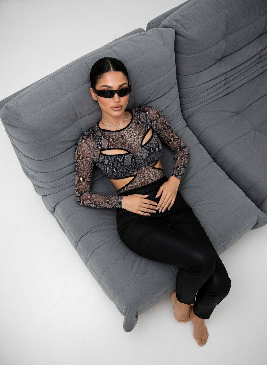 A woman relaxing on a sleek gray sofa, wearing a snakeskin-pattern top and futuristic sunglasses, captured in soft diffused daylight.