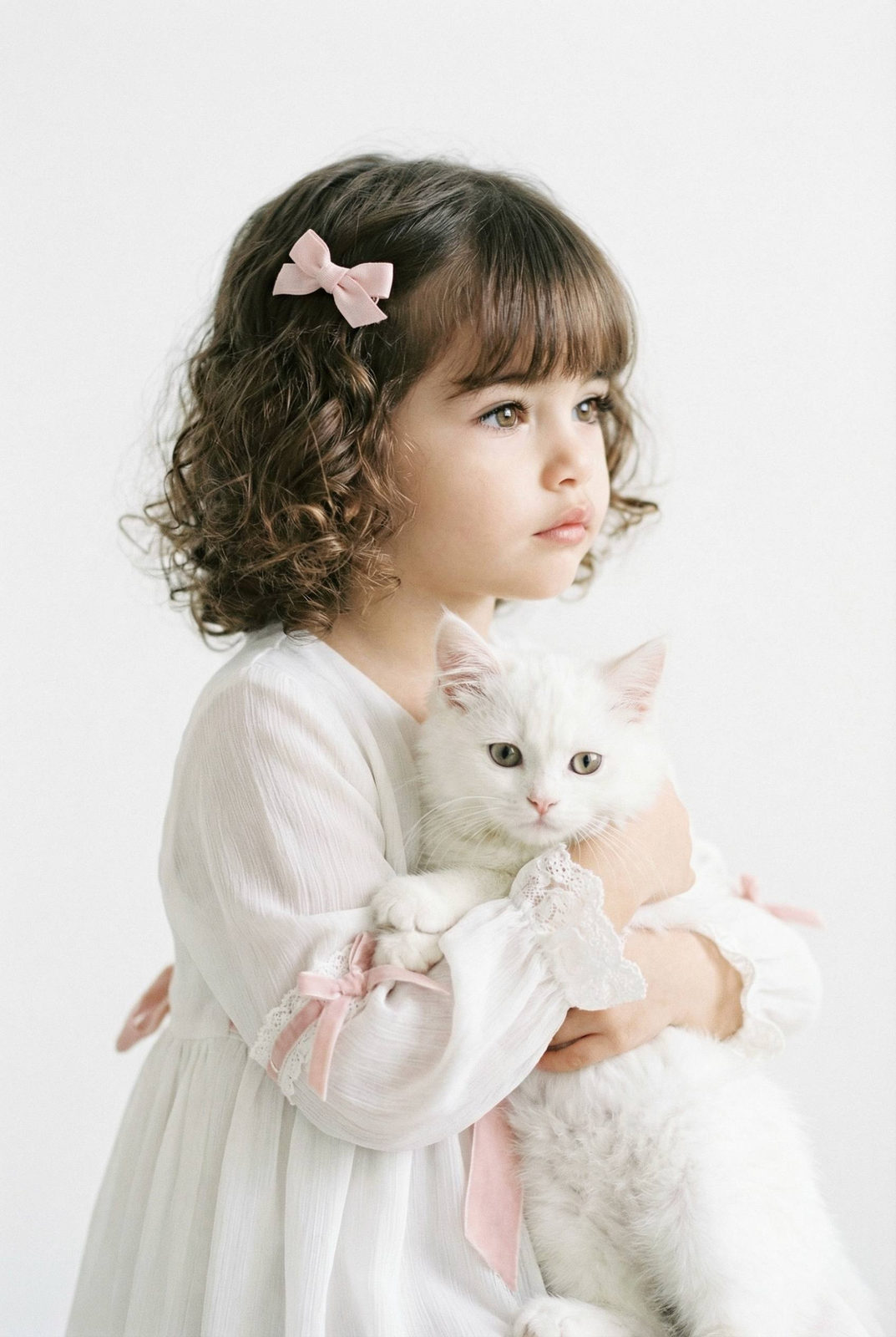 A girl with long brown hair holds a fluffy white cat, wearing a pastel dress with lace cuffs against a soft, minimalistic background.