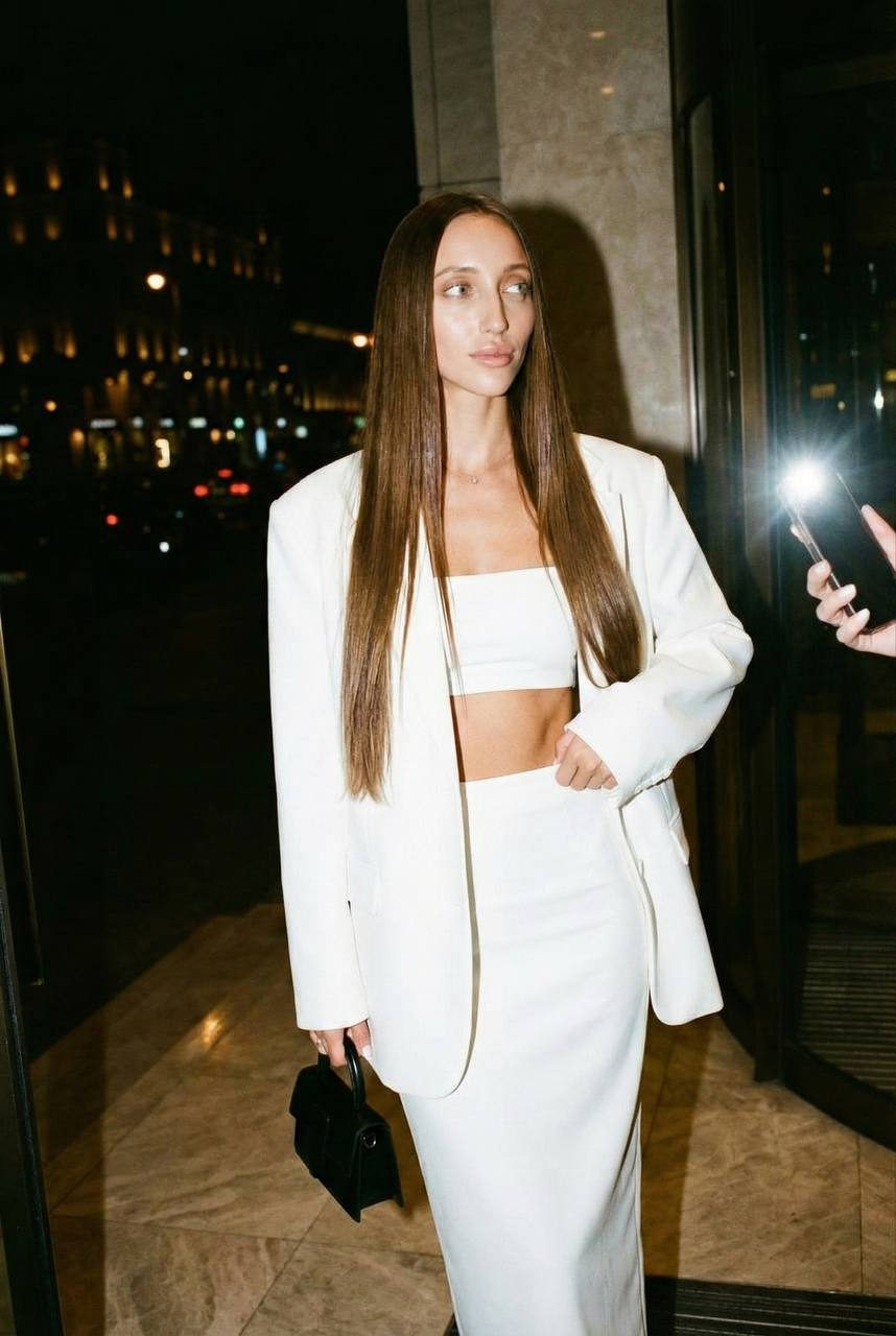 A young woman in a stylish white oversized blazer, standing at a luxurious Monaco hotel entrance with warm city lights in the background.