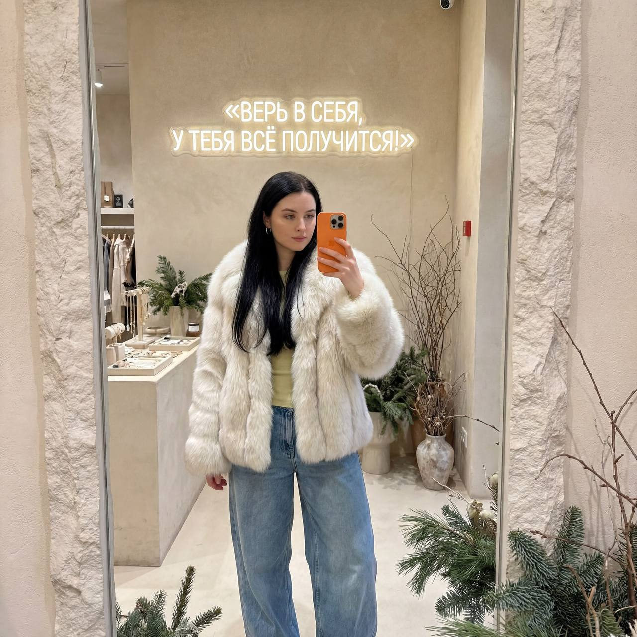 A woman with long straight hair takes a mirror selfie, wearing a fur coat and light top, with a cozy setting and winter decor.