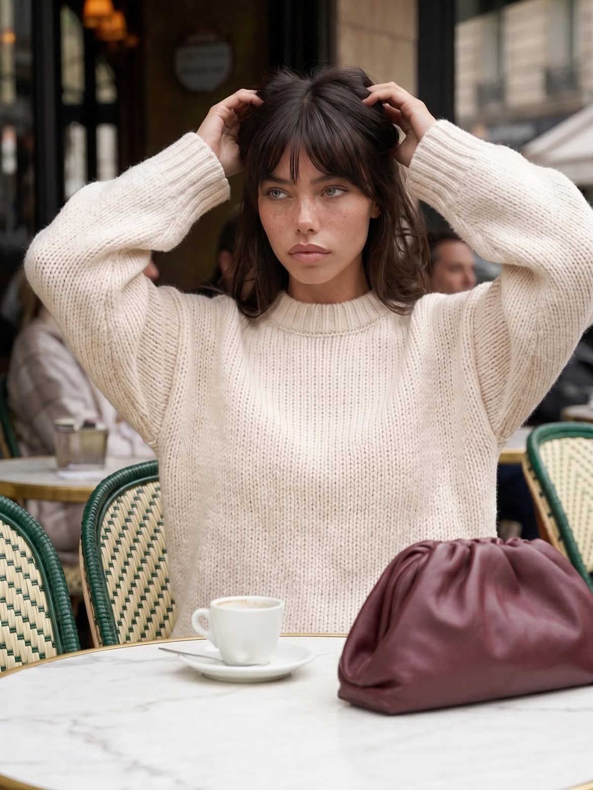 Person in cream ribbed knit sweater sitting at marble cafe table, adjusting hair with a burgundy bag and espresso cup.