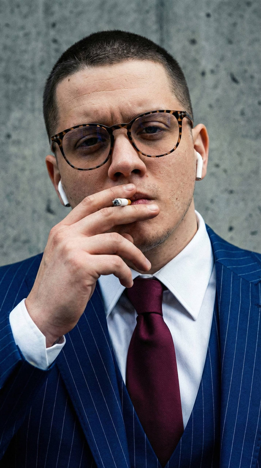 Close-up portrait of a businessman in a blue pinstripe suit, smoking a cigarette with AirPods in a gray concrete background.
