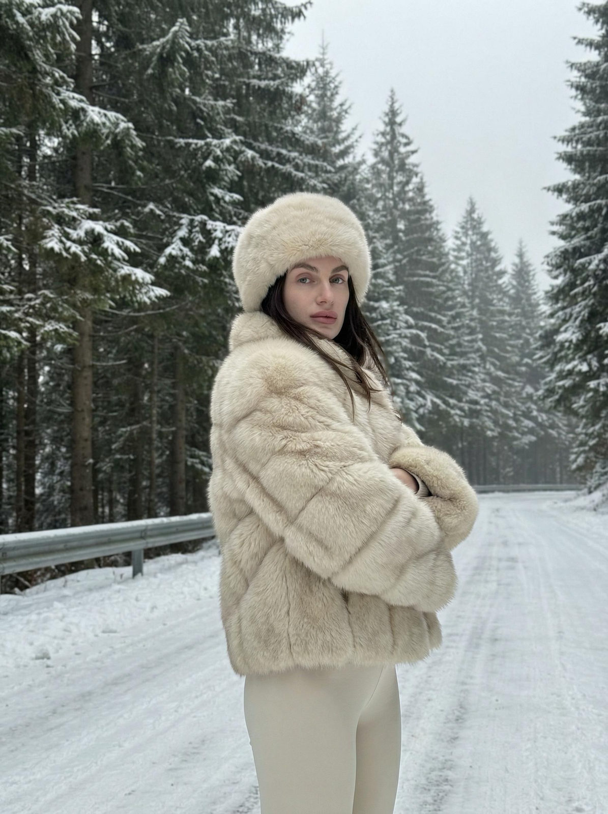 A woman in a cream fur coat stands on a snowy road, surrounded by tall pine trees under a gray sky.