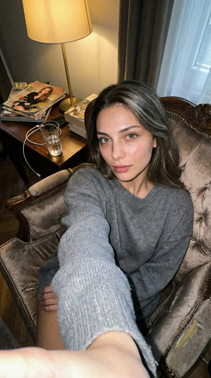 A cozy apartment selfie featuring a subject in soft loungewear sitting on a velvet armchair with scattered personal items in the background.