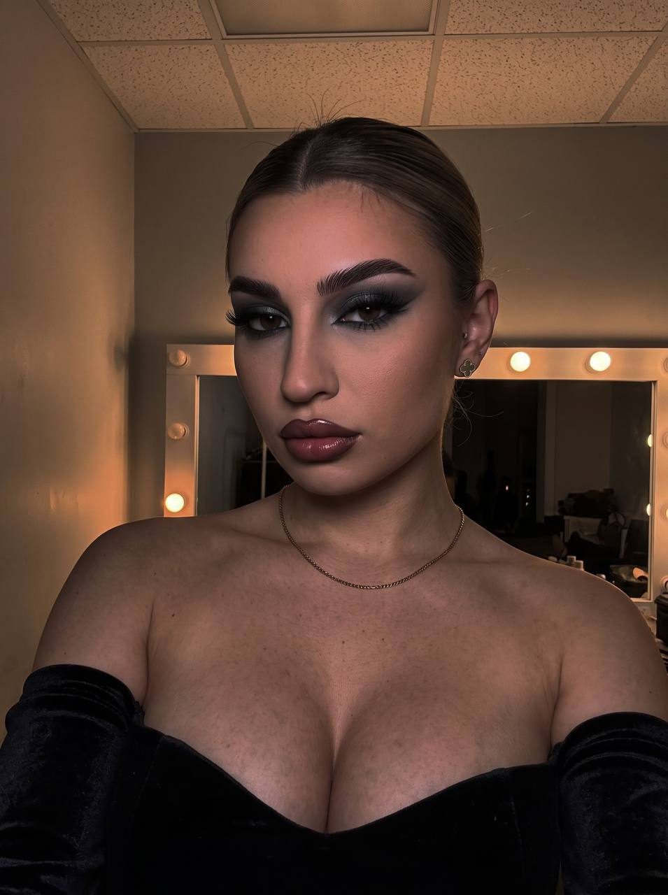 A close-up selfie of a woman with evening makeup featuring smoky gray eyeshadow and glossy dusty-rose lips, wearing a strapless top.