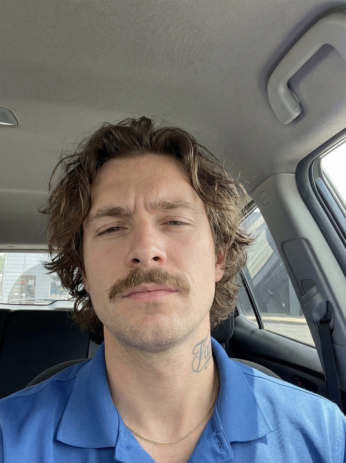 Close-up selfie portrait of a man with wavy hair and a defined mustache, wearing a blue shirt inside a car with natural daylight.