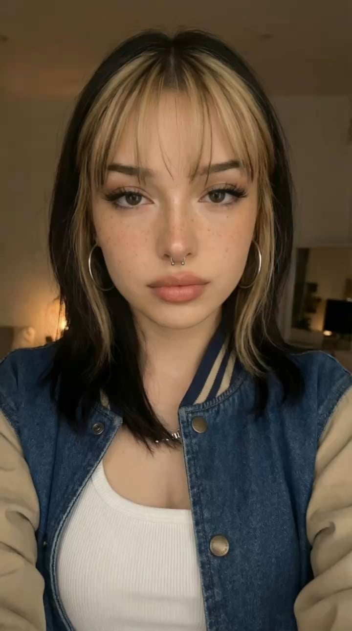 Portrait of a woman in a blue denim varsity jacket with tan sleeves, wearing a white tank top and a silver chain necklace. Soft warm lighting enhances her features.