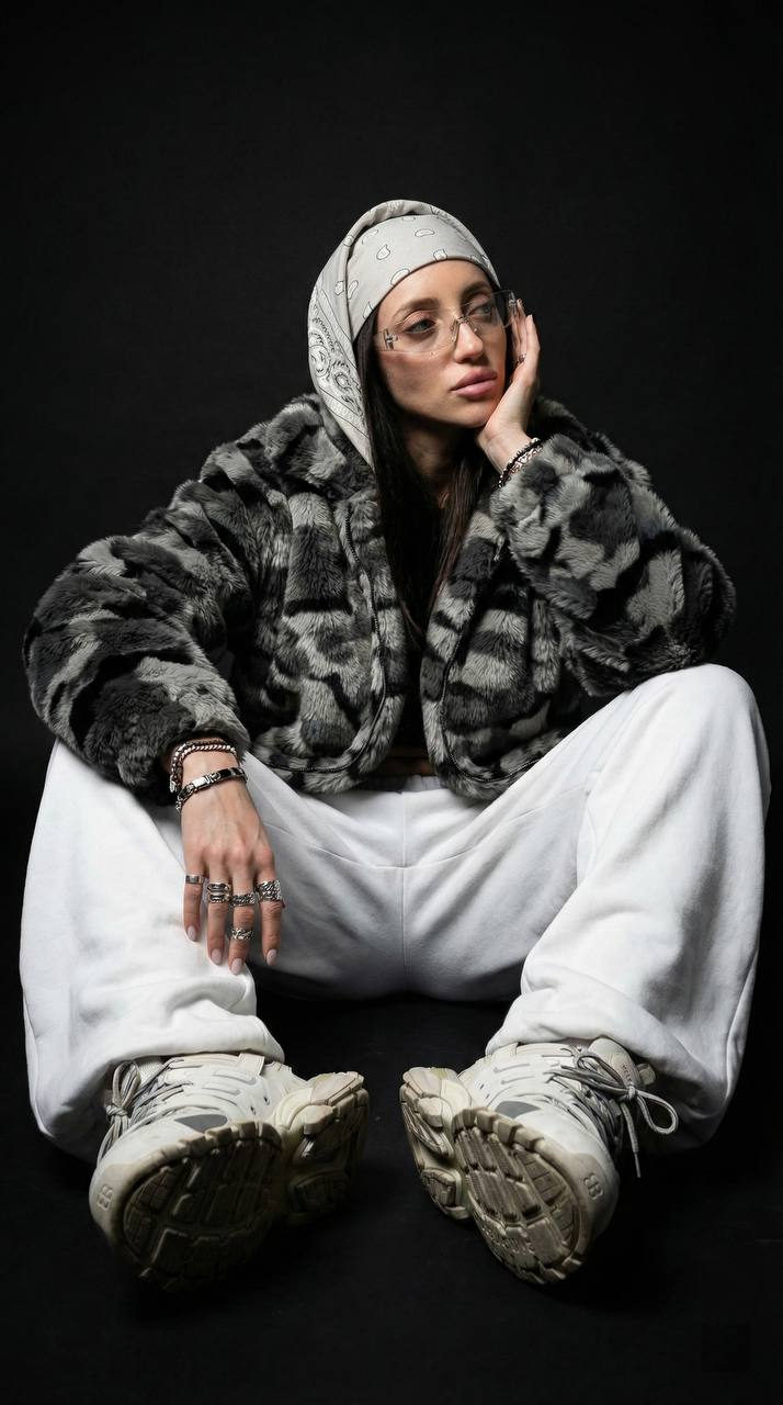 A stylish portrait of a model in streetwear, featuring a fur camouflage jacket, wide sports pants, and massive sneakers against a deep matte background.