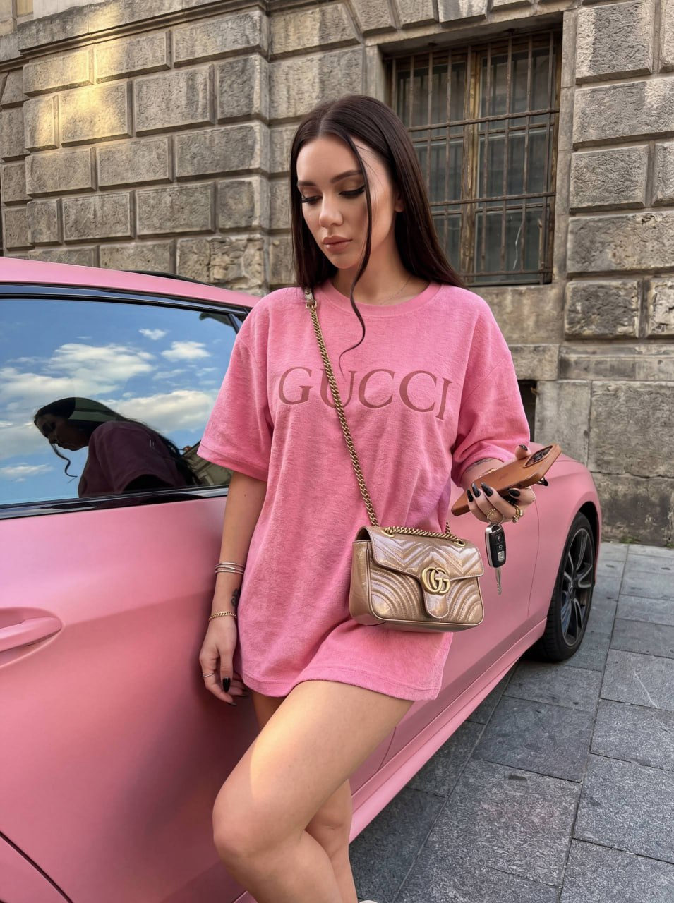 A woman in a pink oversized t-shirt leans against a modern matte-pink car, sunlight highlighting her relaxed pose and stylish accessories.