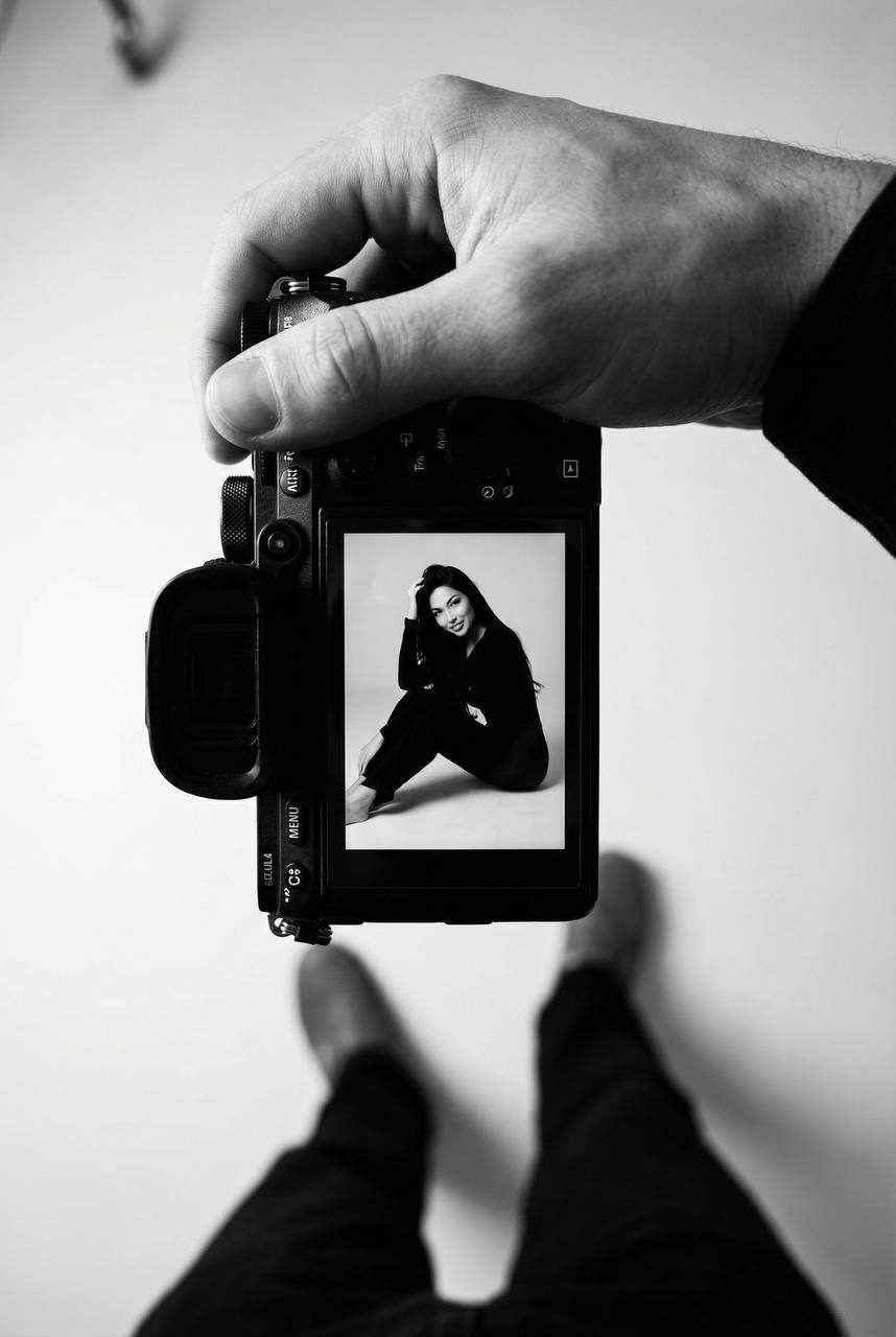 Black and white top-down view of a photographer's hand holding a mirrorless camera with a screen showing a woman smiling in a minimalist studio.