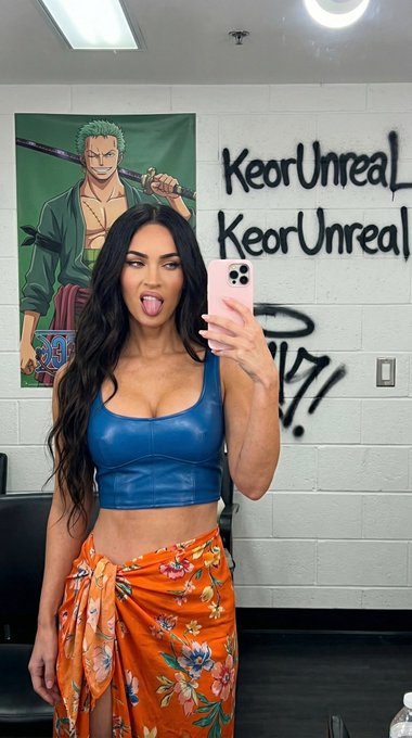 A woman takes a selfie in a dressing room, wearing a blue leather crop top and orange floral sarong, with colorful graffiti in the background.