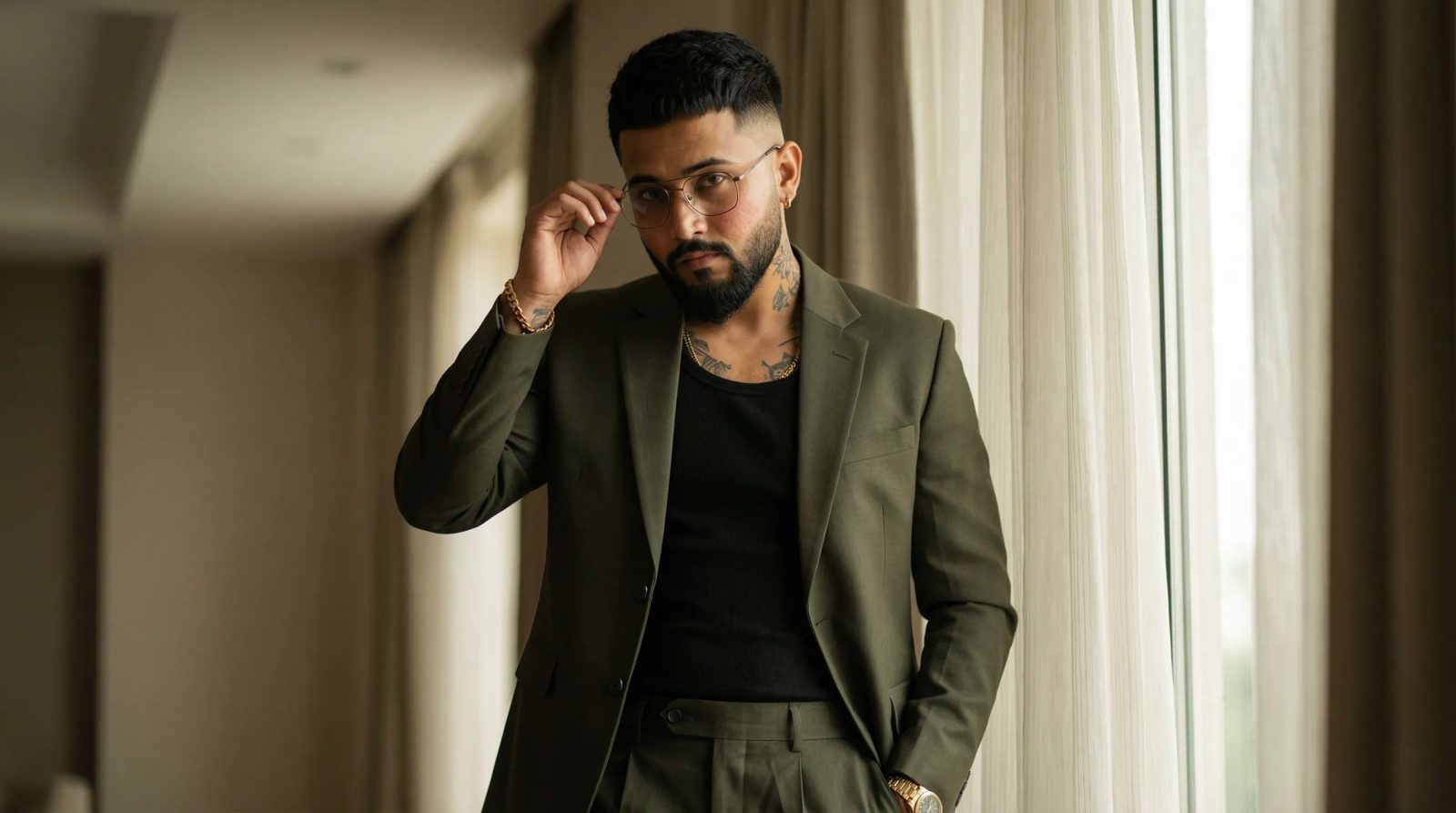 A rugged, masculine man in a dark olive green tailored suit stands beside a window, holding eyeglasses with confidence.