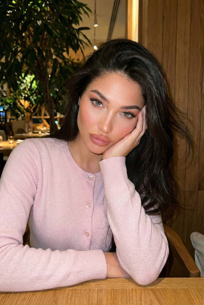 A young woman at a restaurant table in a pastel pink cardigan, looking calmly at the camera with a soft expression.