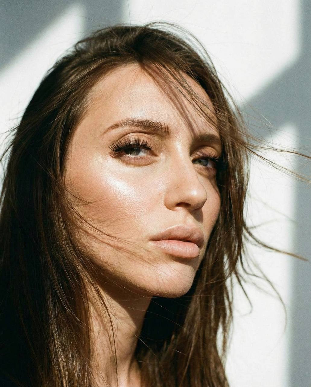 A close-up of a woman's face illuminated by sunlight, with hair fluttering in the wind and emphasizing her expressive eyes.