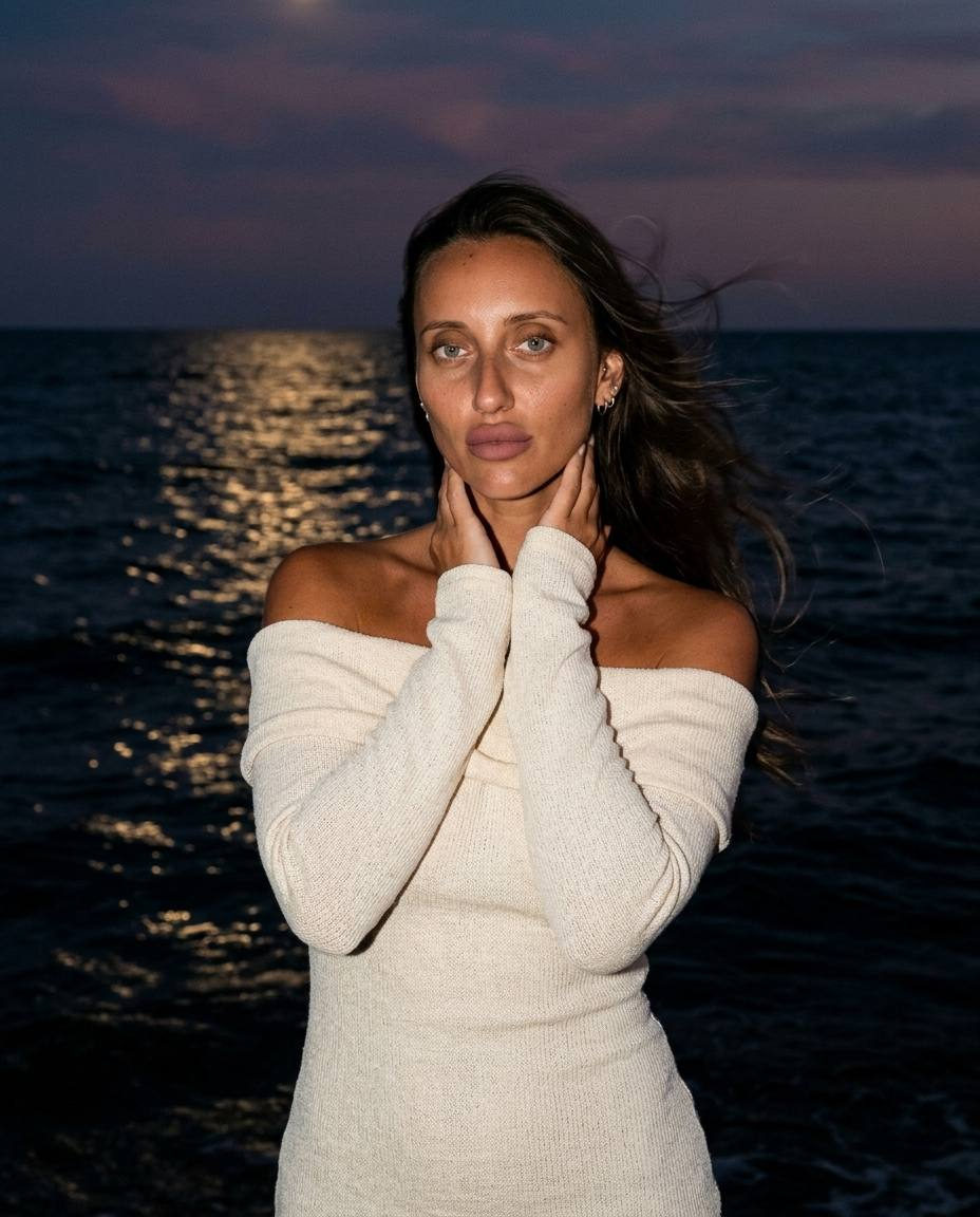 A woman in a light-colored dress stands by shimmering water at night, with golden moonlight reflecting on the waves.