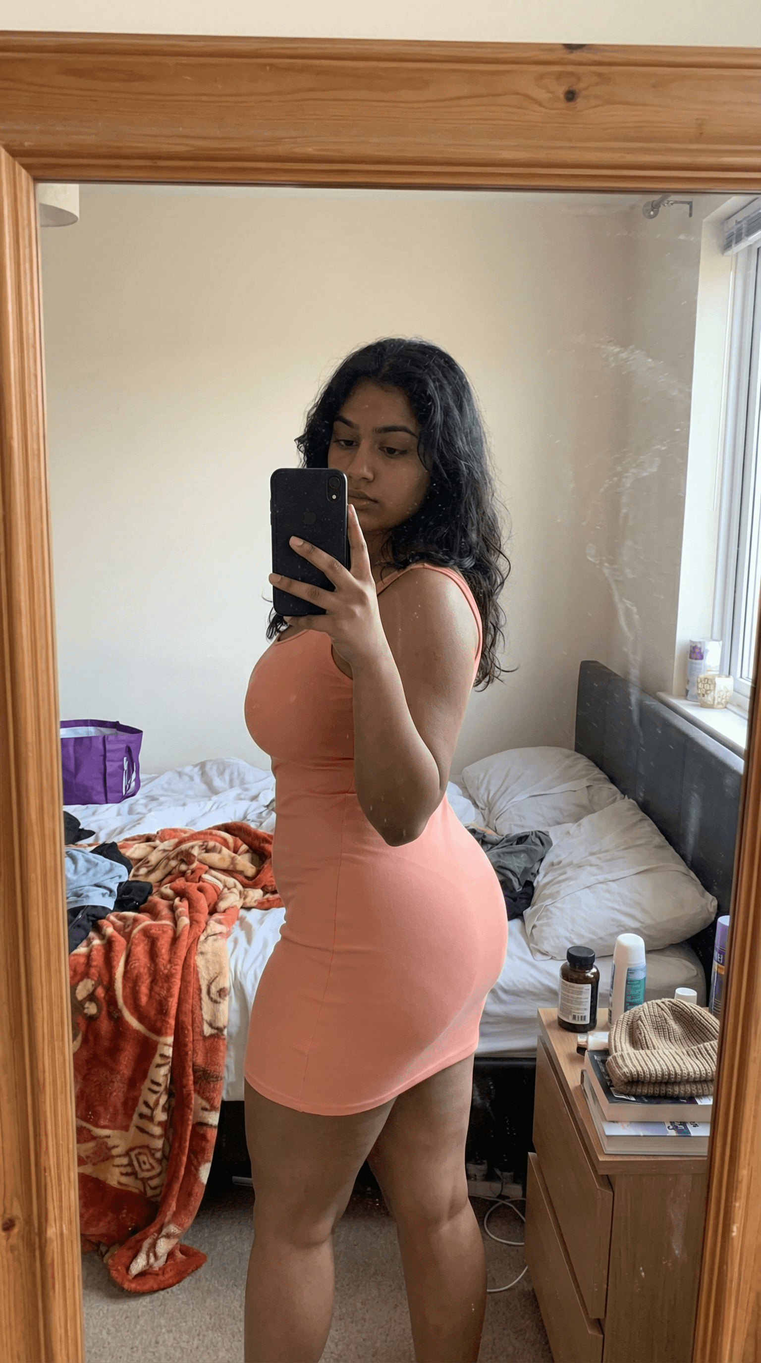 A woman in a peach mini dress takes a mirror selfie in a cozy bedroom with an unmade bed.