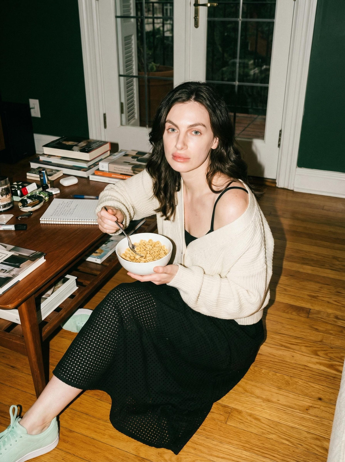 A woman in a cozy, lived-in living room, holding a white bowl of cereal and looking directly at the camera, dressed in a loose cardigan and sneakers.