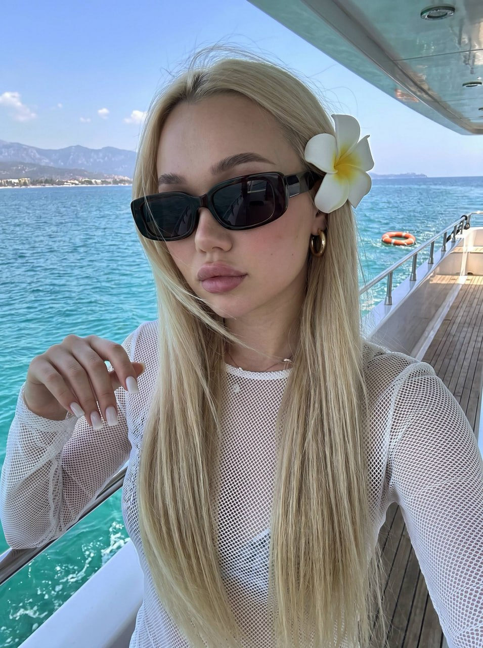 A girl with long straight hair wearing sunglasses, standing on a yacht deck against turquoise water.