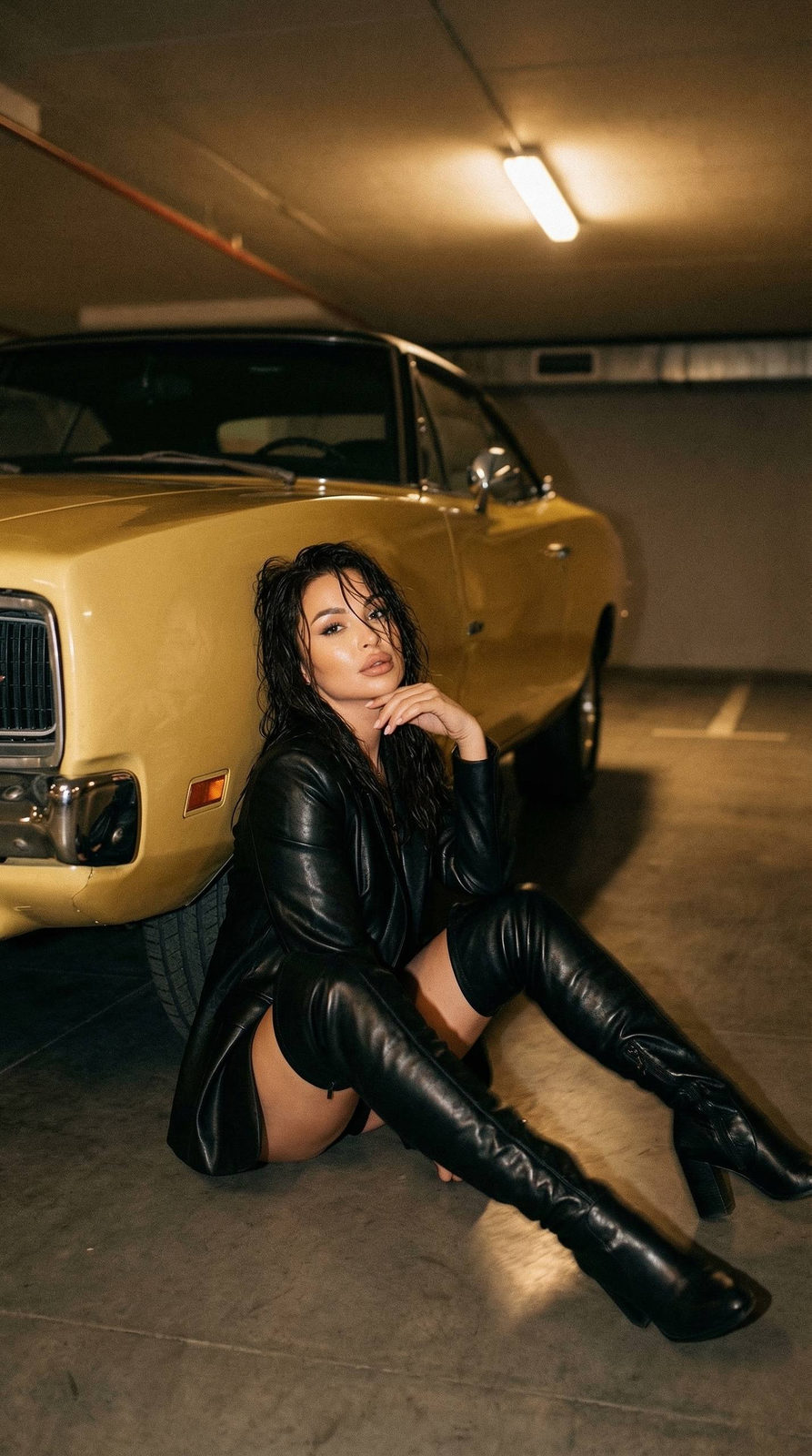 A woman in stylish black leather high boots and a leather jacket, seated on a damp parking lot floor beside a retro yellow Dodge Charger, with soft warm lighting.