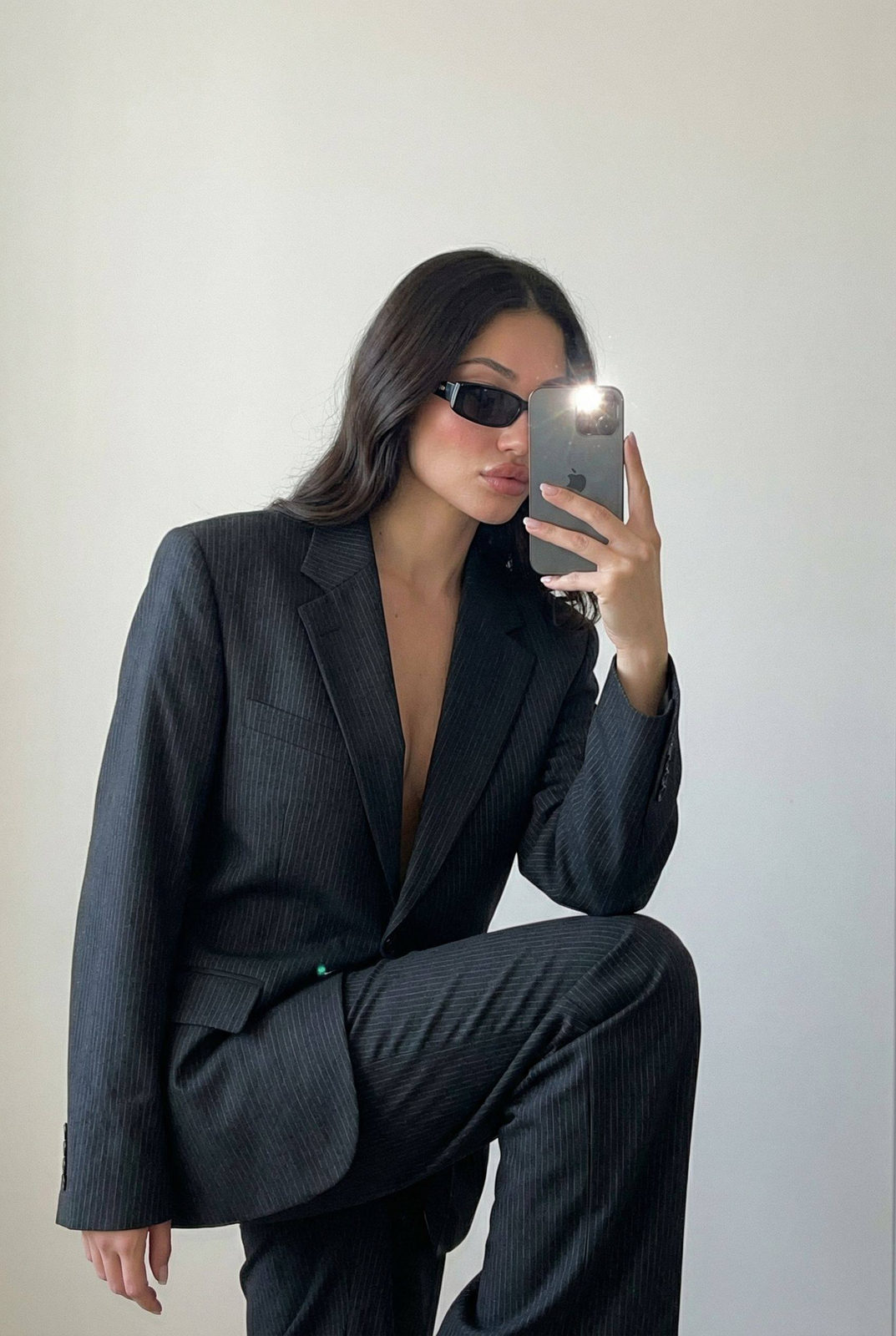 Slender woman in pinstripe suit taking a mirror selfie indoors, posing confidently against a minimal light-colored wall.