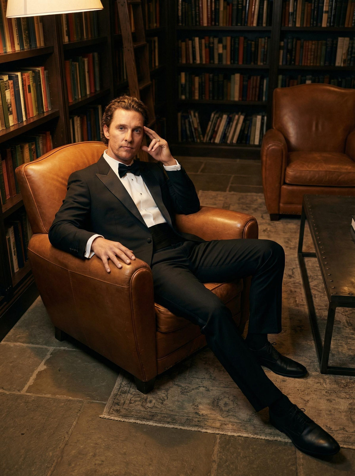 A tuxedo man reclining in a leather chair in a vintage library, with relaxed posture and intense gaze.