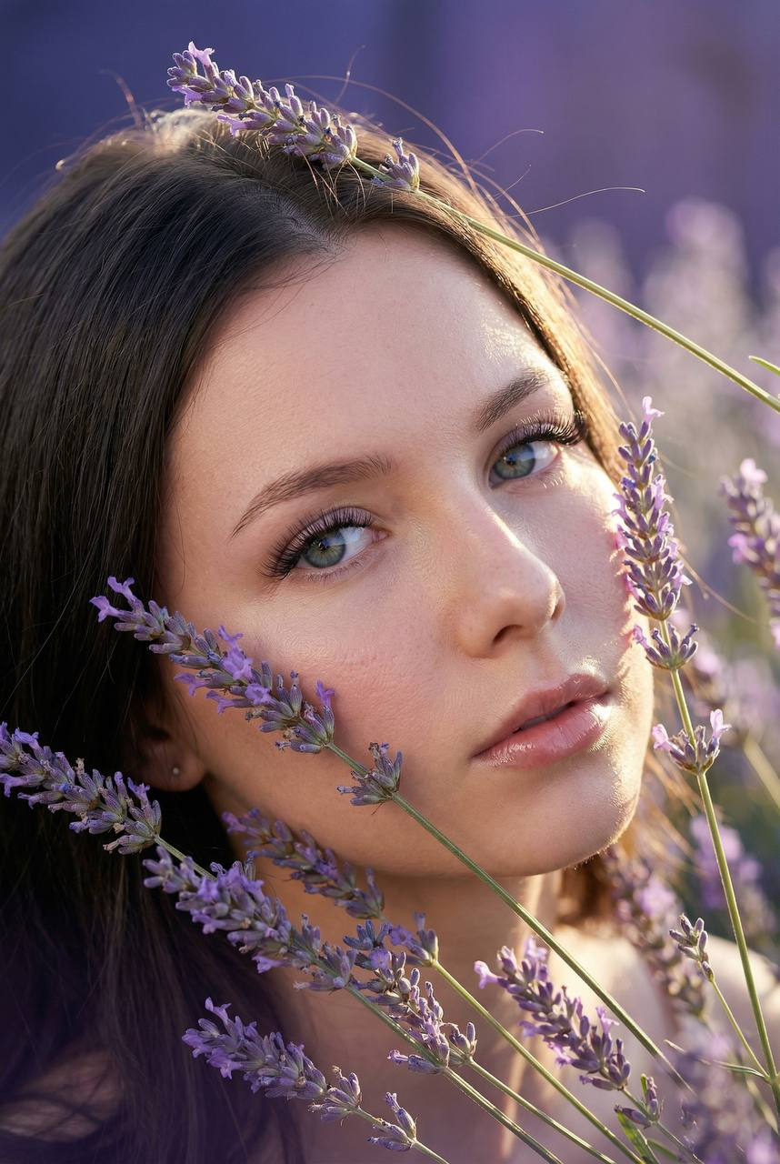 Close-up macro portrait of a person in a lavender field, with delicate inflorescences framing the face and soft lavender branches brushing the cheek.