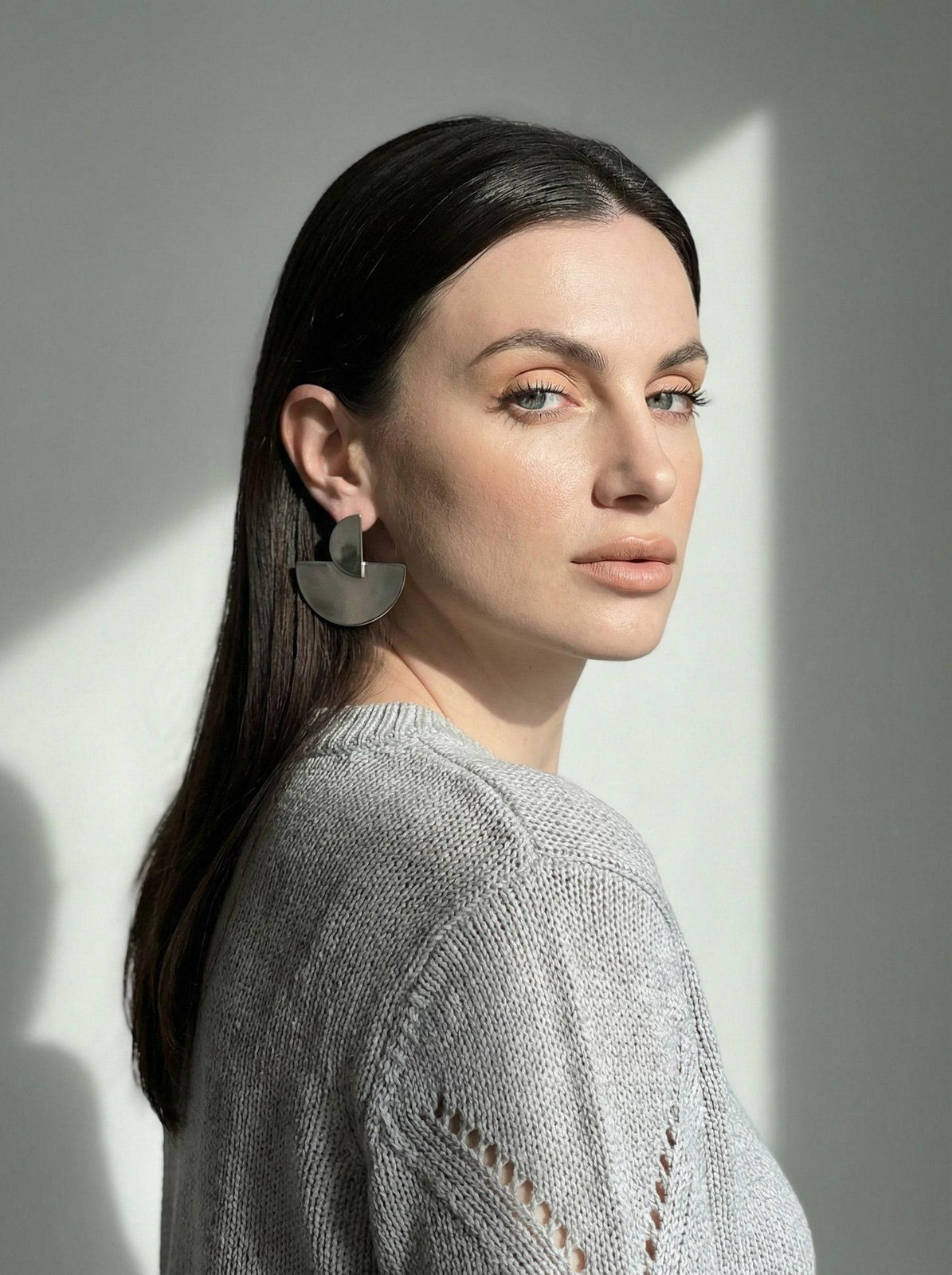 Studio portrait of a woman against a light gray wall, showcasing detailed makeup and a textured gray jumper.