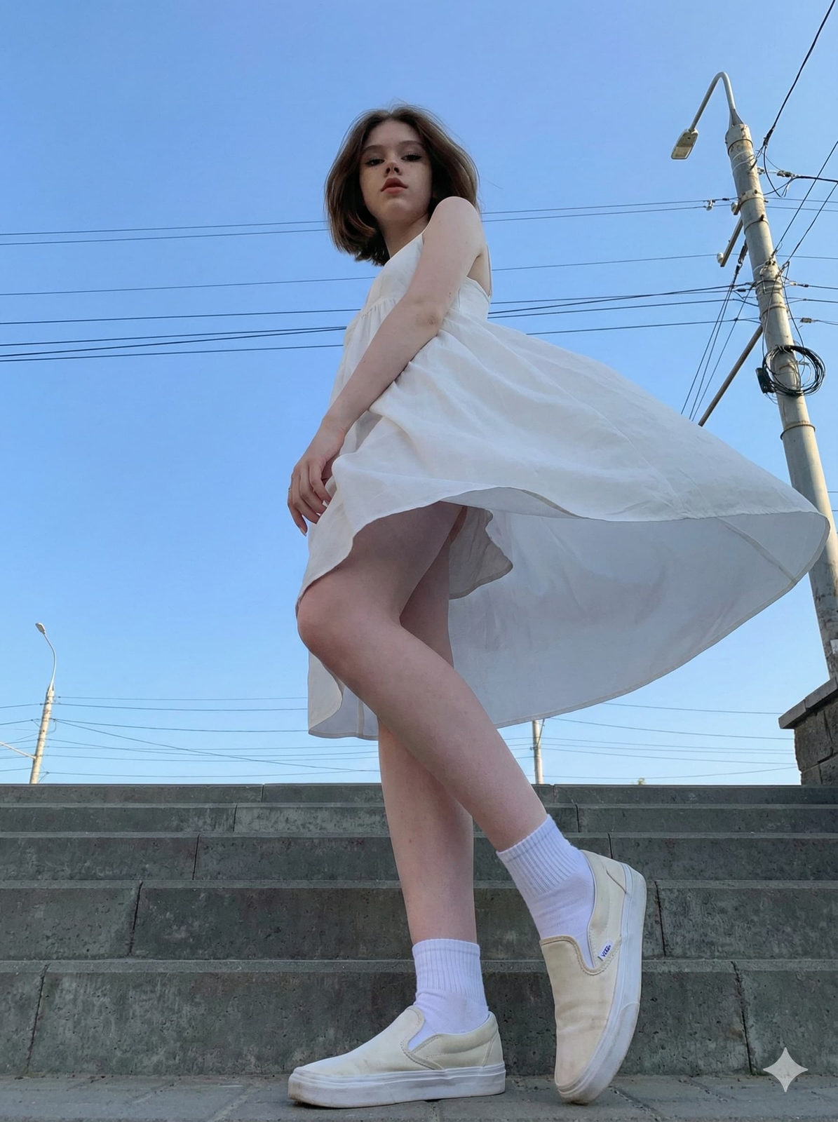 A woman in a flowing dress stands confidently at the bottom of a staircase, viewed from a low angle against a clear blue sky.