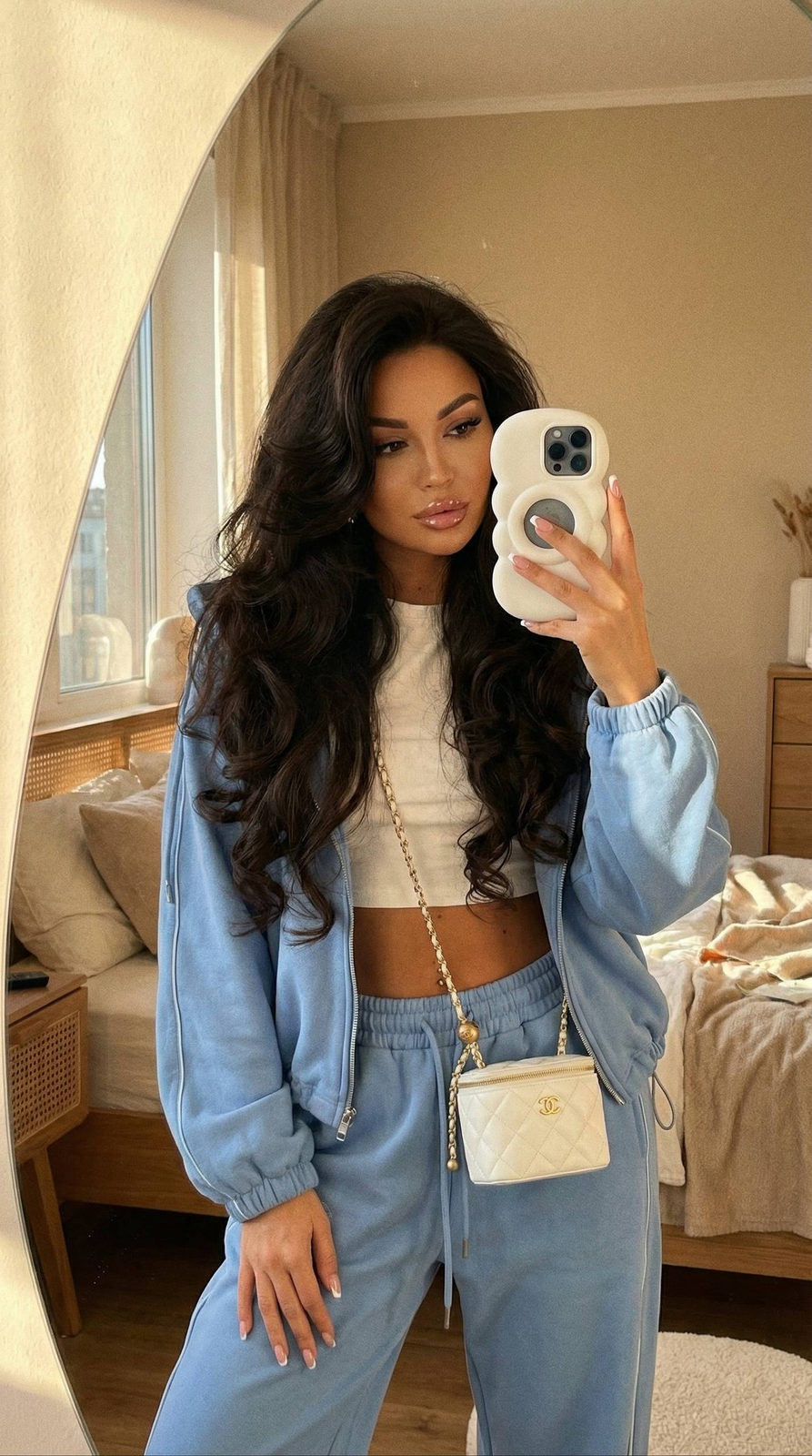 A girl taking a selfie in a frameless mirror, showcasing glossy lips and voluminous, curled hair in a warm-toned bedroom.