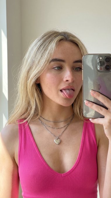 Close-up selfie of a woman wearing a pink tank top with a smartphone, playful expression captured in soft light and shadows.